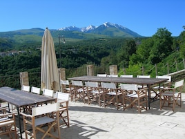 Un área de comedor al aire libre con suelo de piedra y mobiliario de madera, con vistas a un paisaje montañoso con picos nevados y vegetación exuberante.