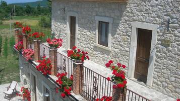 A stone building with a balcony adorned with red flowers, a wooden door, and a window with a shutter.