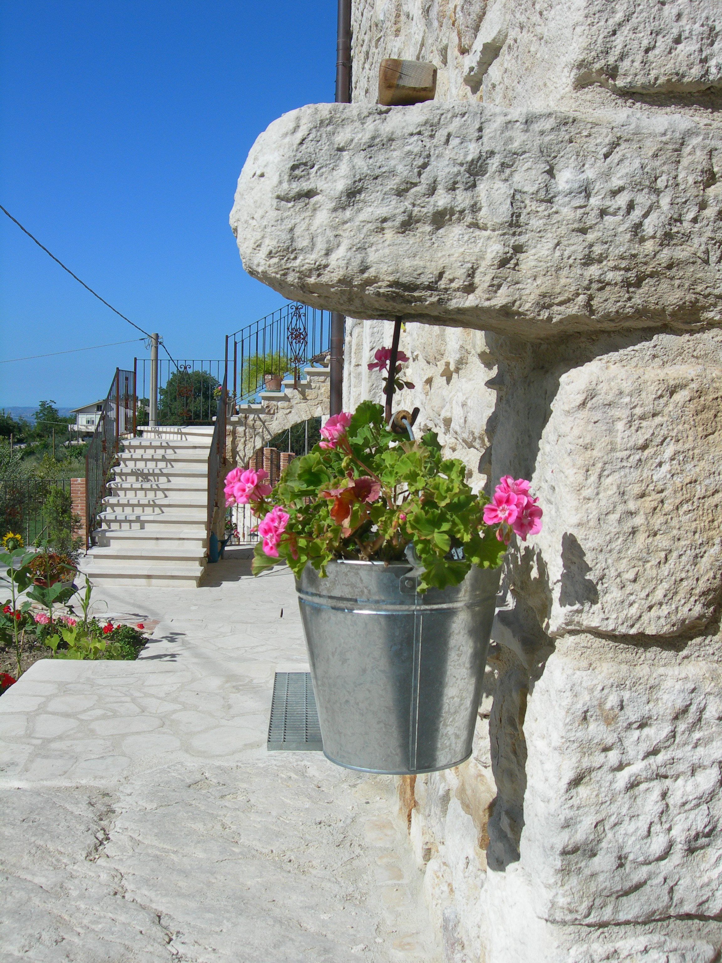A stone building with a metal bucket planter filled with pink flowers hanging from a ledge.