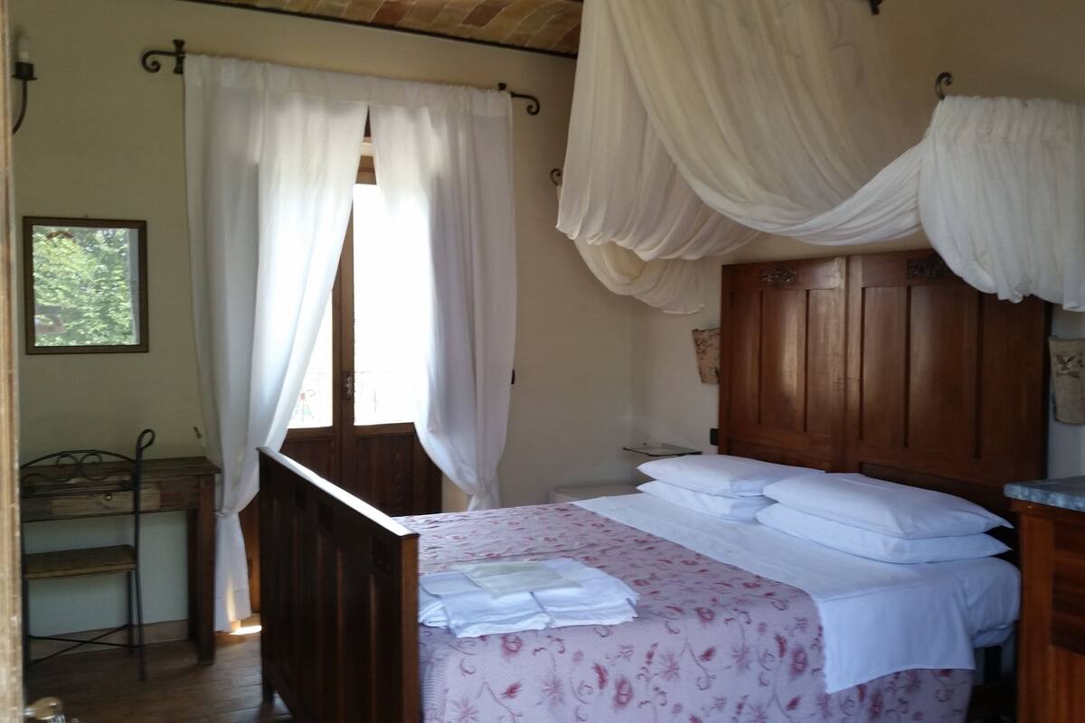 Een slaapkamer met een houten bed, witte gordijnen en een raam met uitzicht op groen.