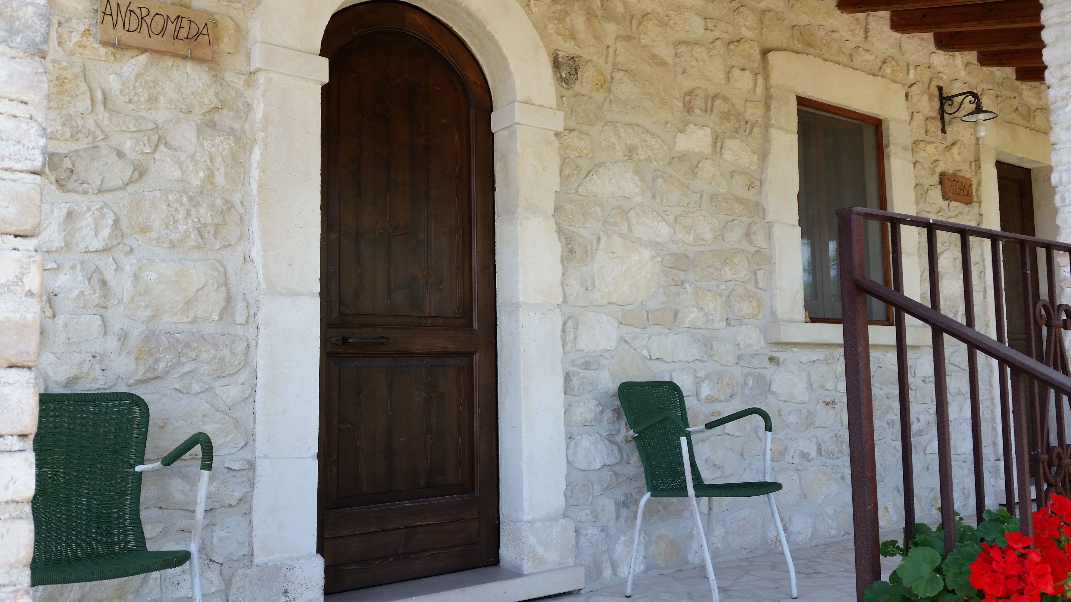 Un bâtiment en pierre avec une grande porte en bois, deux chaises en osier, une rambarde en métal et une enseigne sur laquelle on peut lire « ANDROMÈDE ».