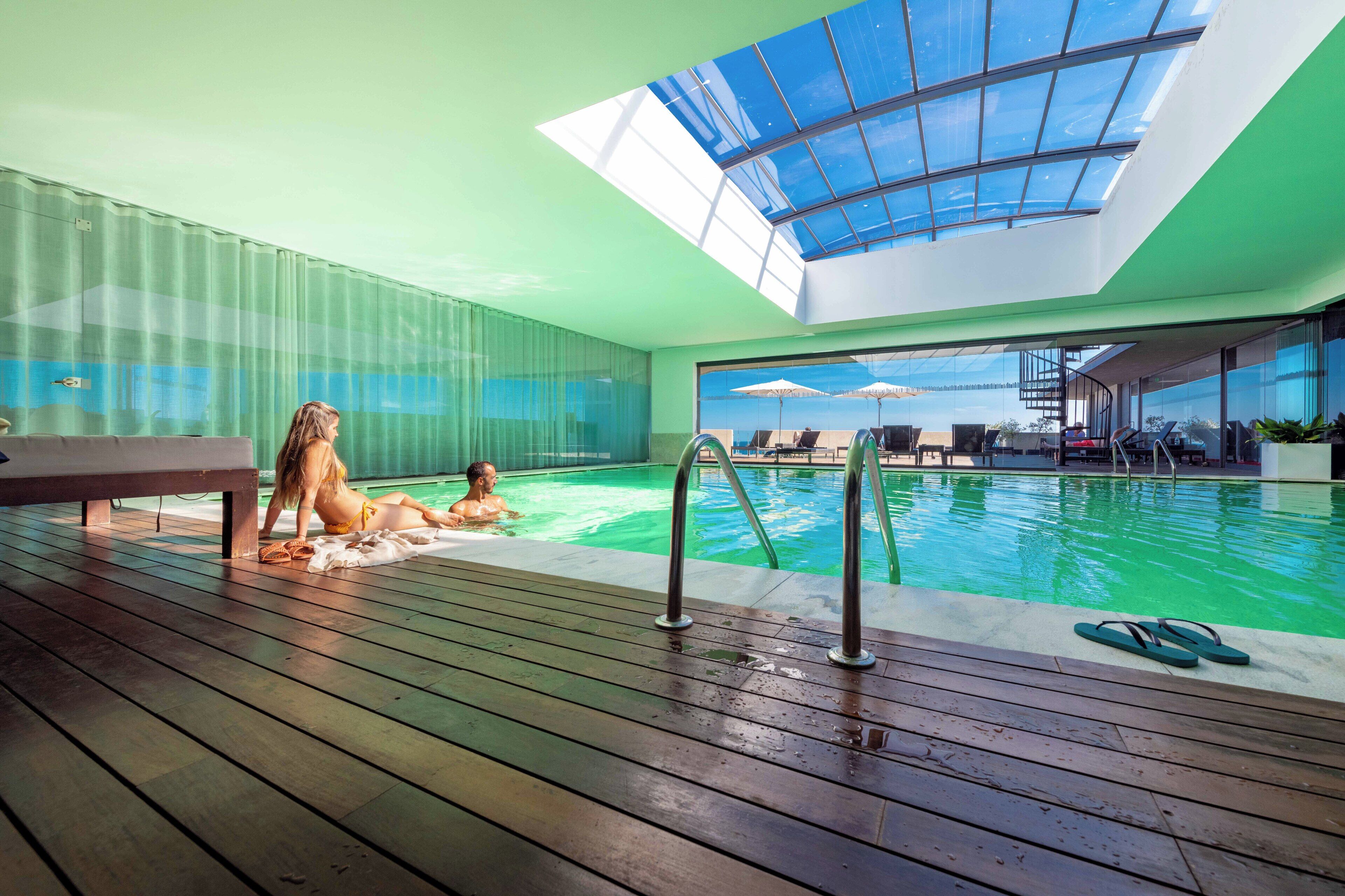 A modern indoor swimming pool with a skylight, wooden deck, and a view of the city.