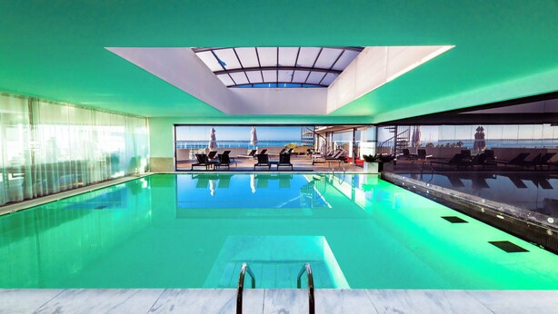 A modern indoor swimming pool with clear glass walls, a view of the sea, and a sunlit ceiling.