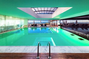 A modern indoor swimming pool with clear glass walls, a view of the sea, and a sunlit ceiling. - Pestana Cidadela Cascais - Pousada & Art District (Cascais)