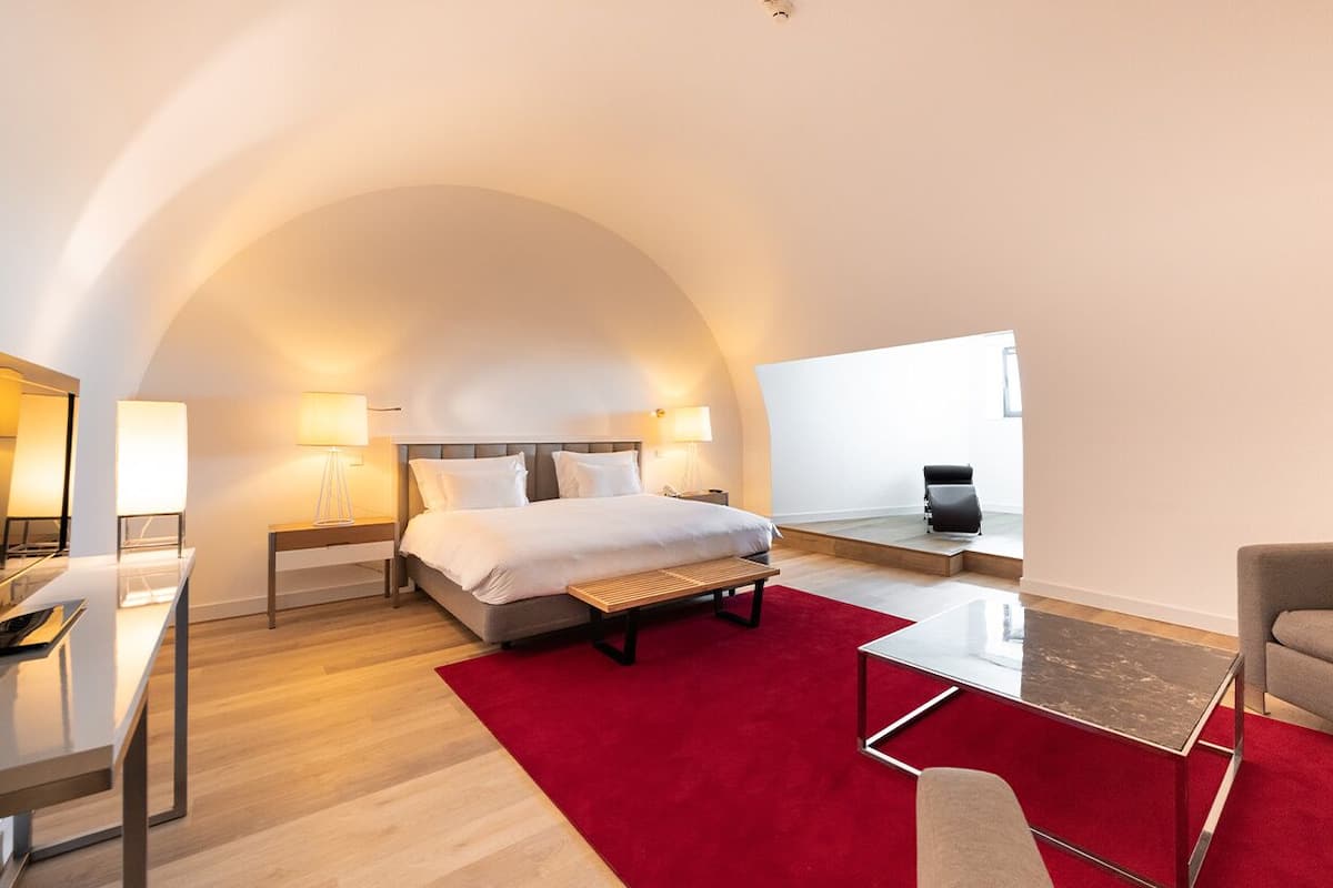 Una habitación de hotel moderna con techo curvo, una cama con ropa de cama blanca, alfombra roja, una mesa de centro de vidrio y un sofá.