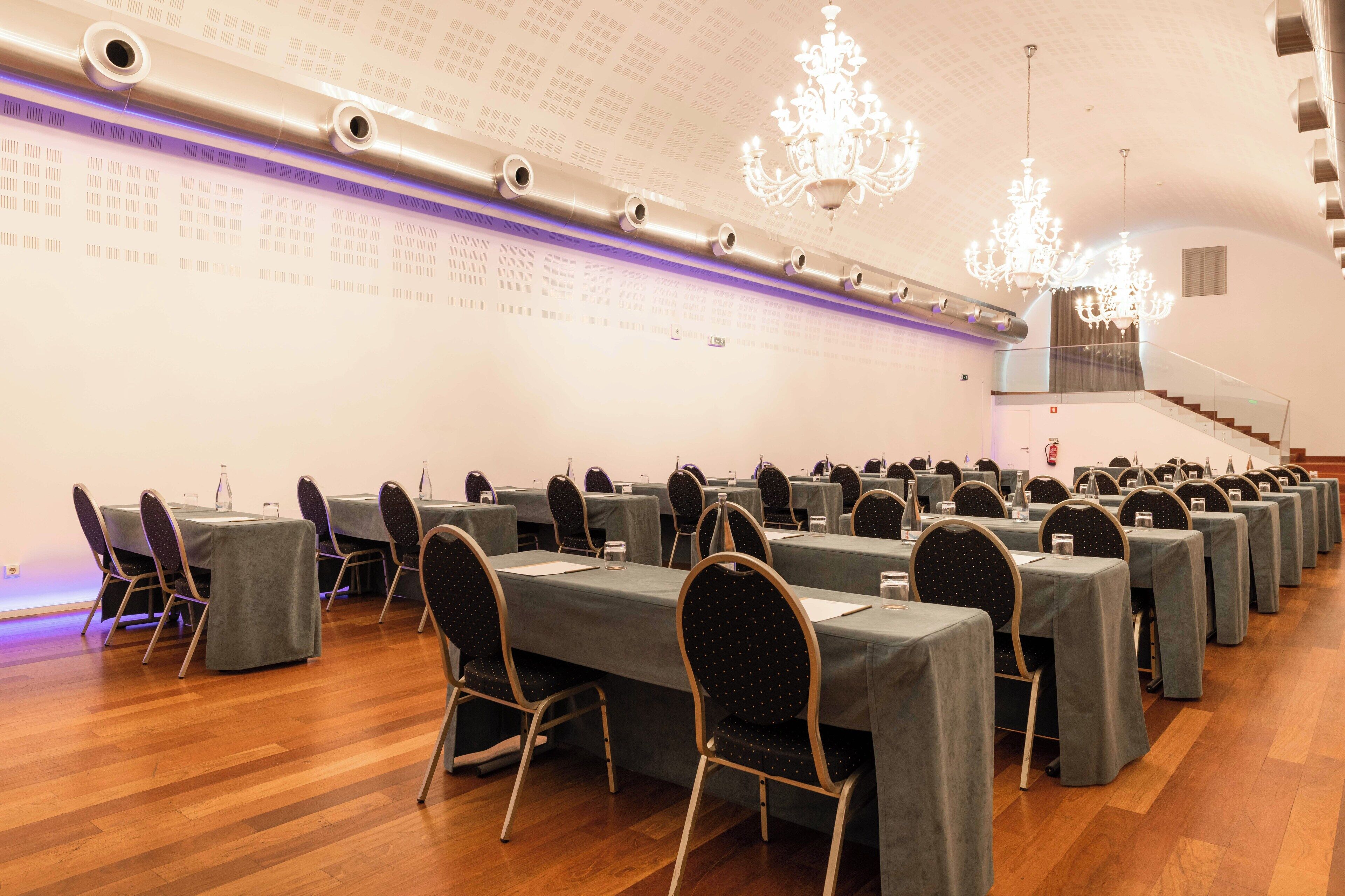 Un salón de conferencias con filas de mesas y sillas, candelabros y una escalera.