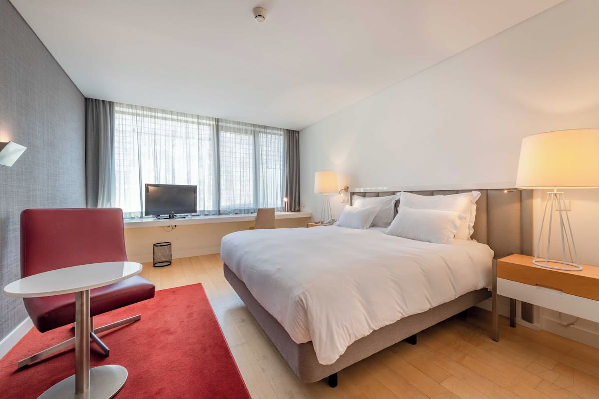 Habitación de hotel con una cama grande, un sillón rojo, una mesita, televisión y ventana con cortinas.