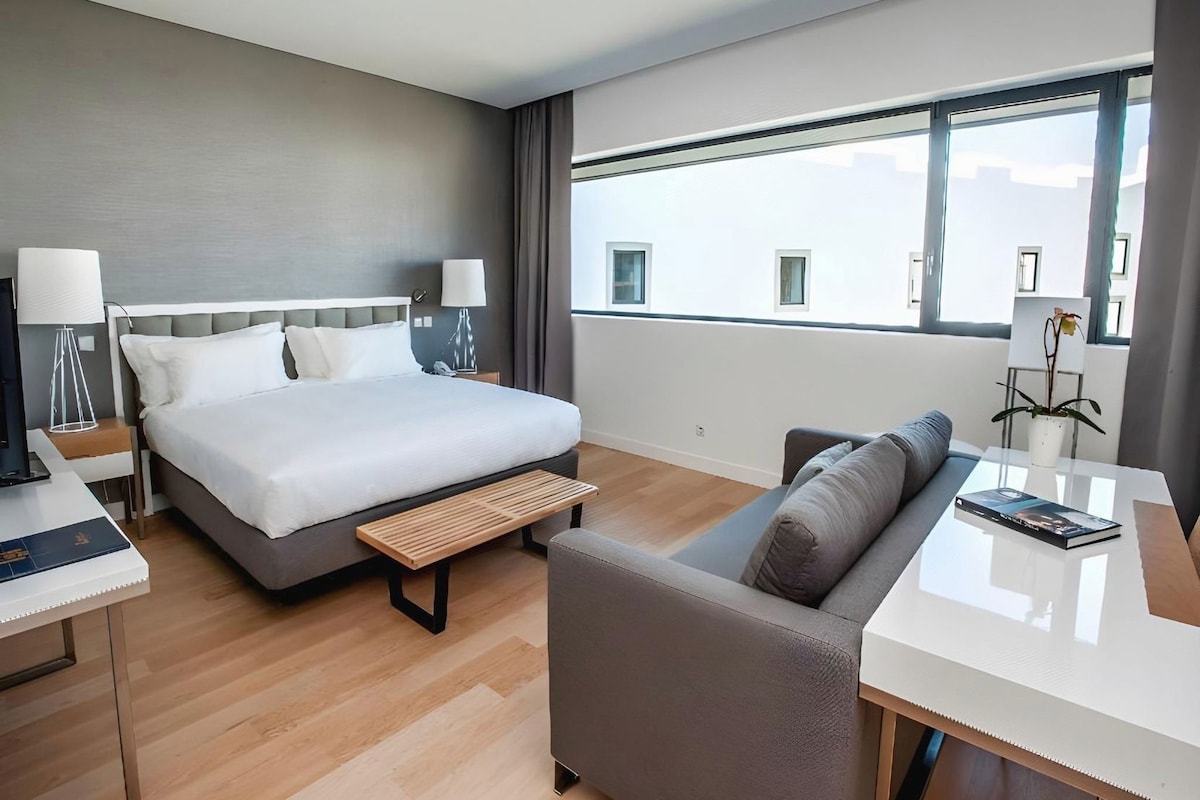 Habitación de hotel con cama, sofá, una mesita y ventana con vistas a edificios.