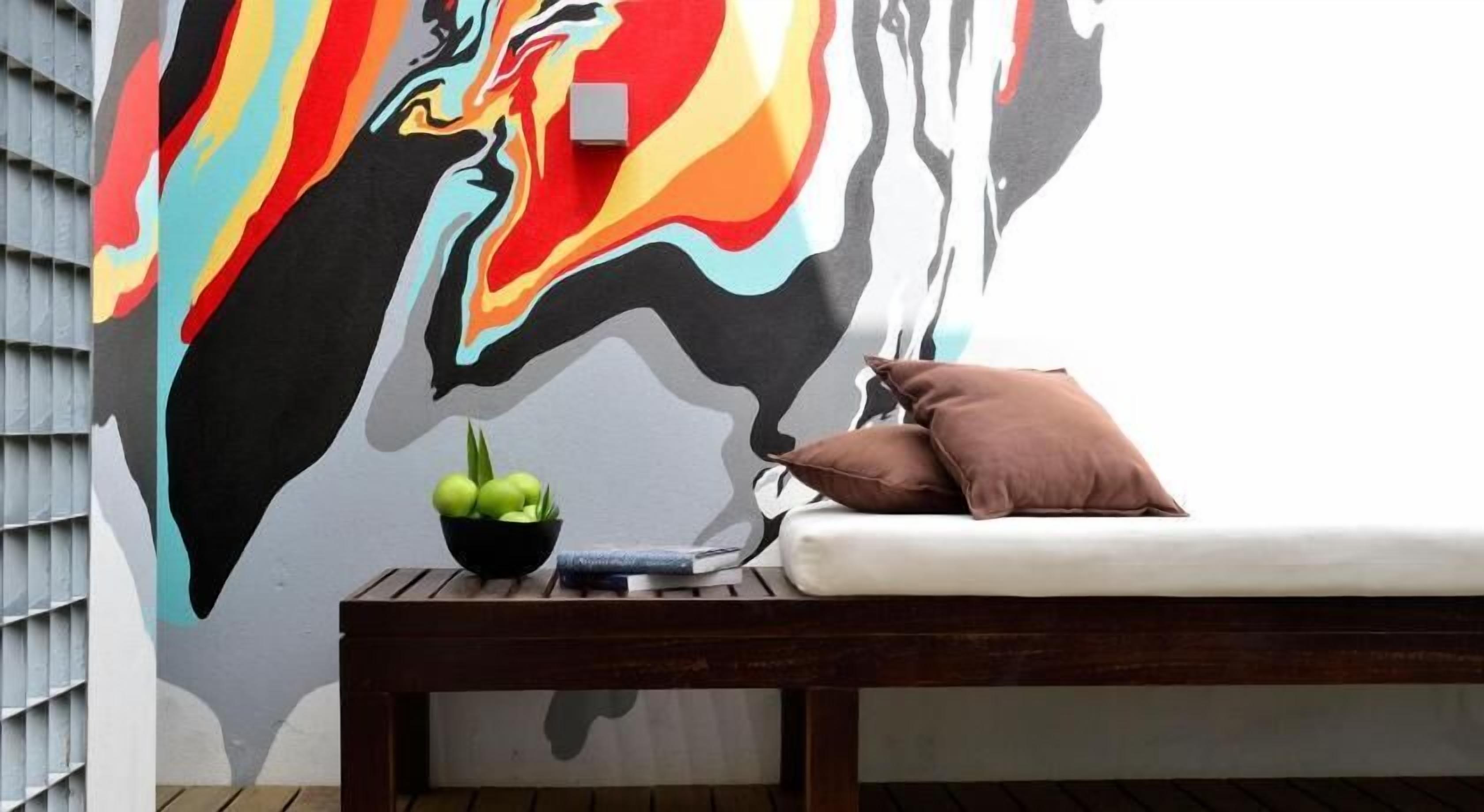 Une pièce décorée d’une fresque murale, avec un banc en bois, un panier de fruits et un coussin.