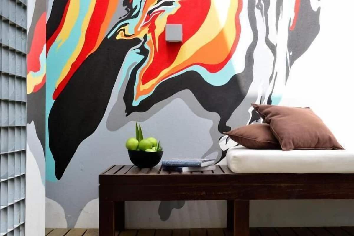 Une pièce décorée d’une fresque murale, avec un banc en bois, un panier de fruits et un coussin.
