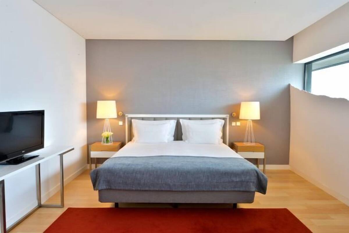 Habitación de hotel moderna con una cama grande, lámparas de noche, un televisor de pantalla plana y una ventana.