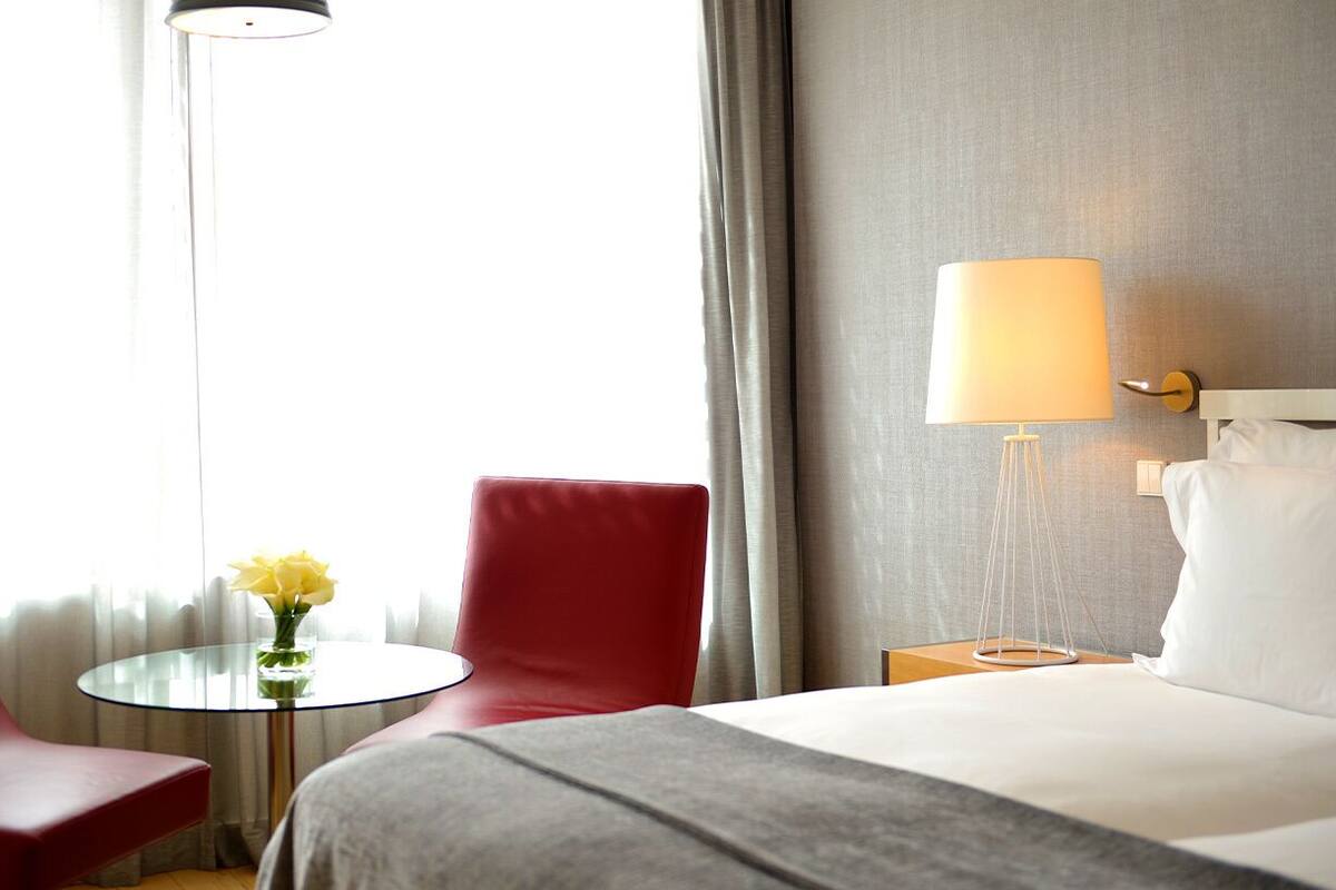 Una habitación de hotel con una cama, una silla roja, una mesita redonda con un jarrón de flores amarillas, una lámpara de noche y vistas al exterior a través de cortinas translúcidas.