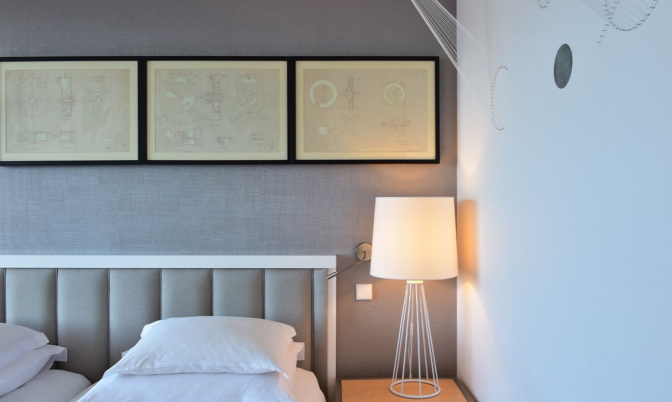 Una habitación de hotel con una cama, una mesita de noche, una lámpara y tres dibujos arquitectónicos enmarcados en la pared.