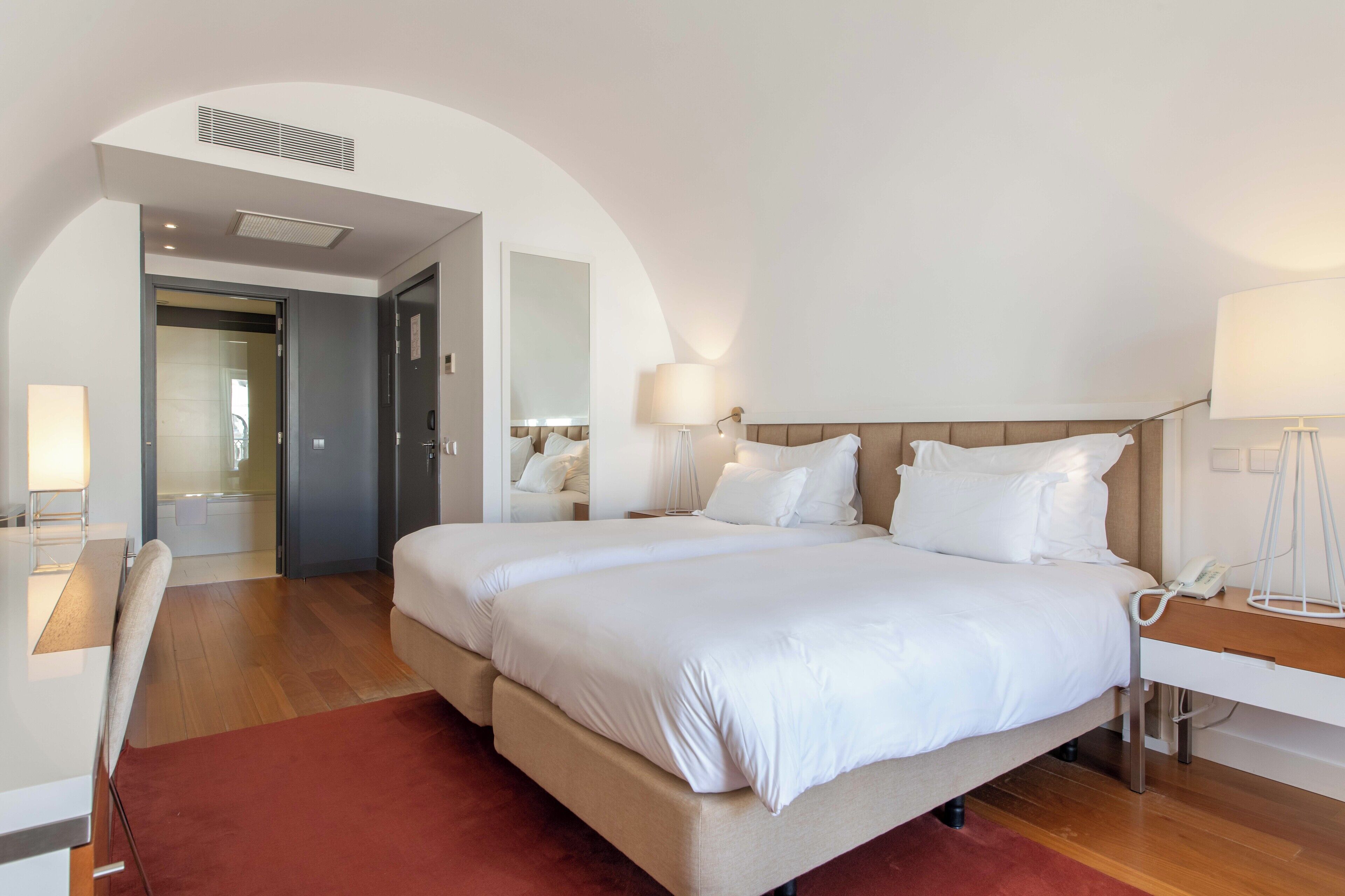 A modern hotel room with a large bed, a desk, and a chair. There is a mirror on the wall and a door leading to the bathroom.