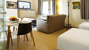 A hotel room with a bed, a sofa, a dining table with chairs, a TV, and a coat rack. - Apartments Ypres (Ypres)