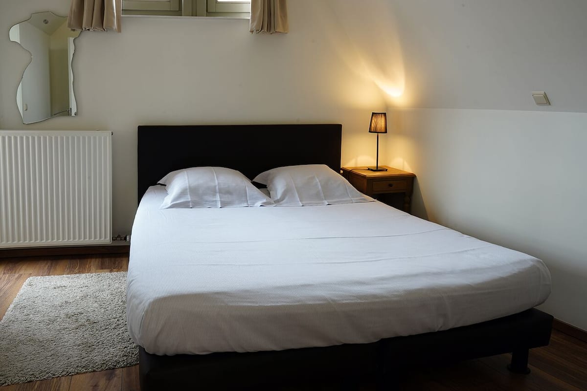 A neatly made bed with white linens, a bedside lamp, and a radiator on the wall.