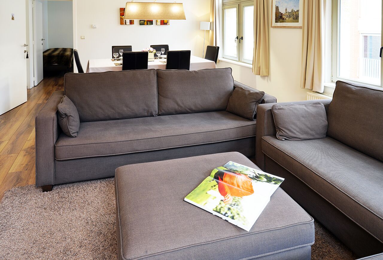 Un salon moderne avec un canapé gris, un pouf assorti et un coin repas à l’arrière-plan.
