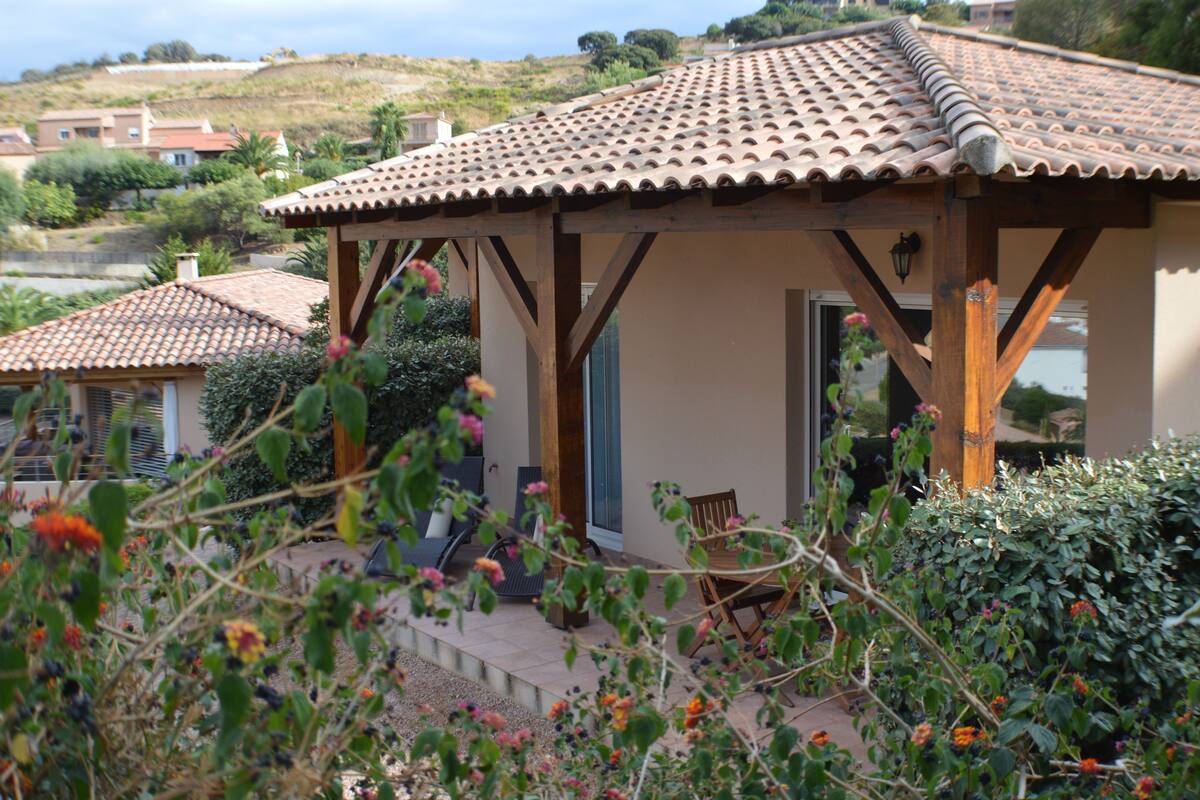 Una casa con techo de tejas, un porche cubierto con mesa y sillas, y un jardín con diversas plantas y flores.
