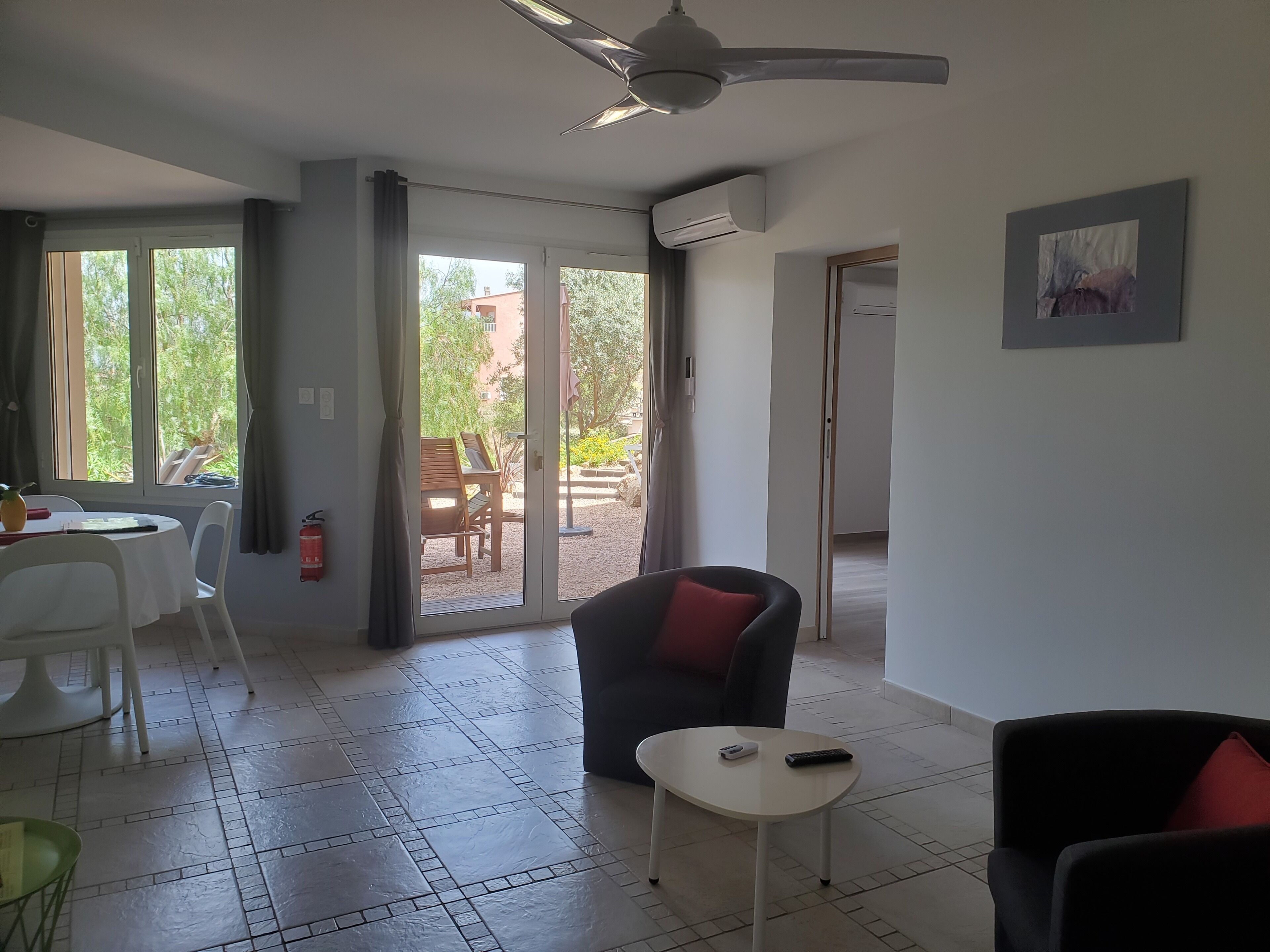 Une pièce dotée d’un ventilateur de plafond, d’une table à manger avec des chaises, d’un canapé, d’une petite table avec une télécommande et d’une porte donnant sur l’extérieur.