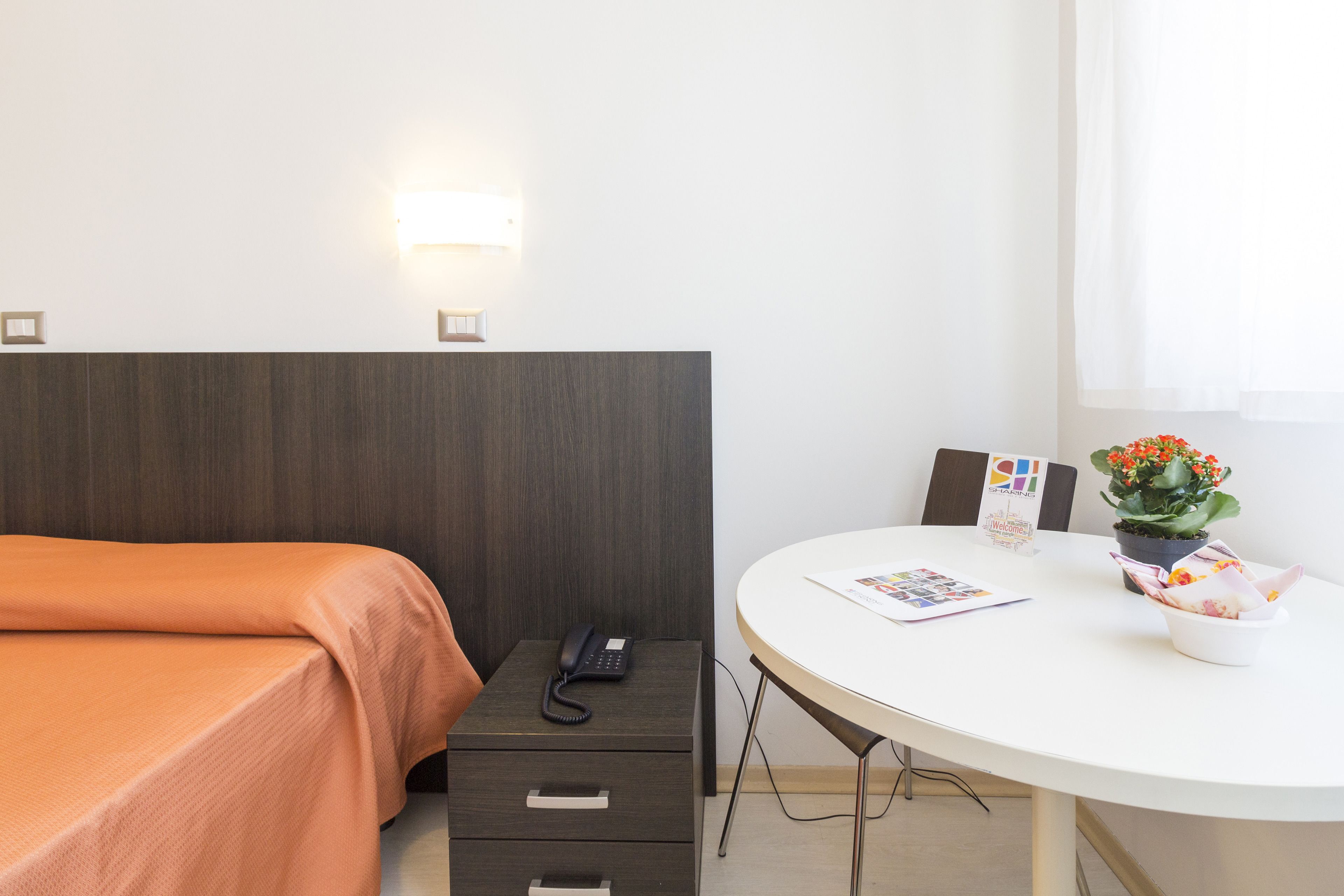 Habitación de hotel con cama, mesita de noche con teléfono, una pequeña mesa con una planta y un frutero, y una lámpara fijada a la pared.