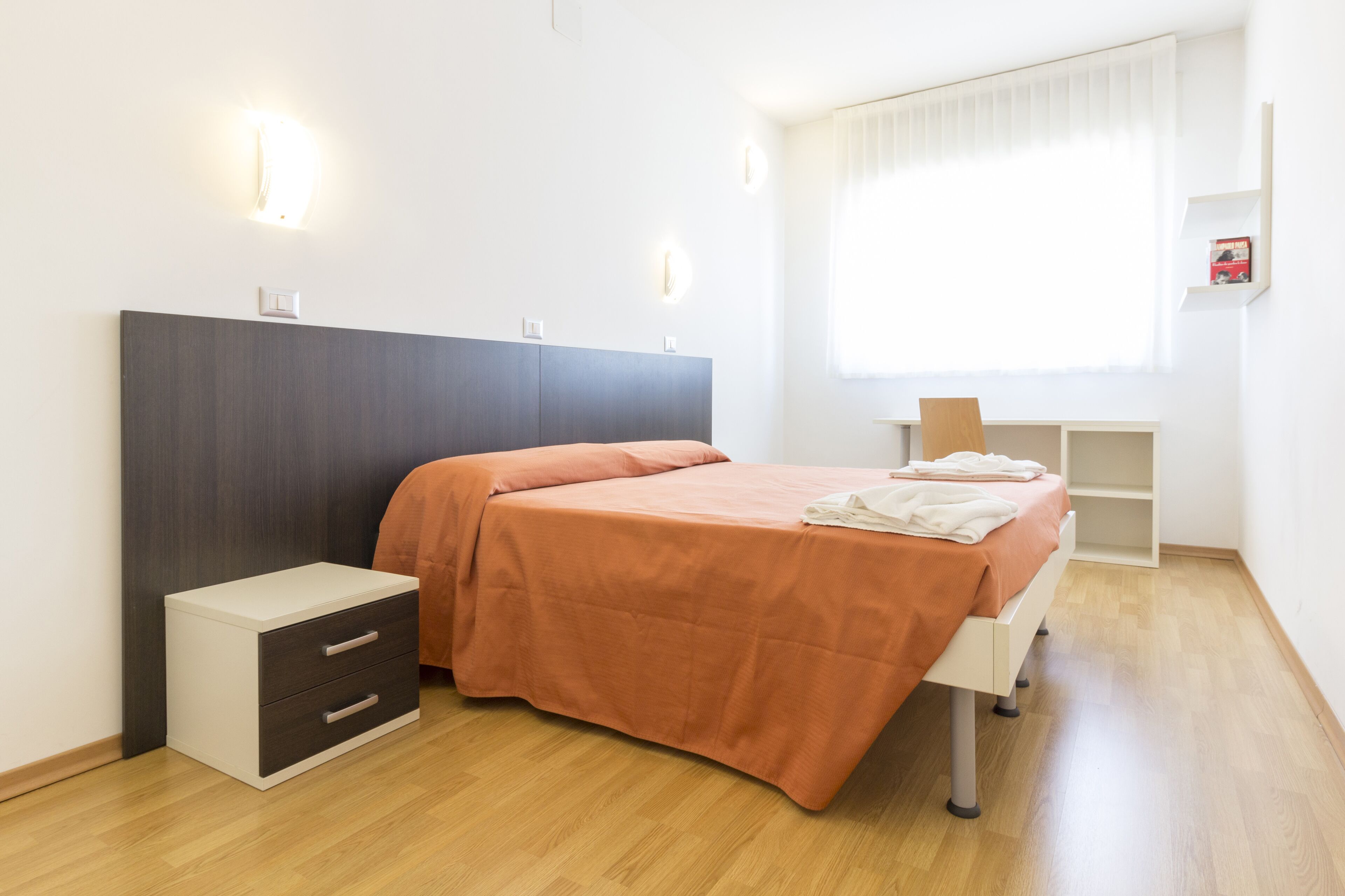 Ein Schlafzimmer mit Bett, Nachttischschubladen, einem Nachttisch, einem Schreibtisch und einem Fenster mit Jalousien.