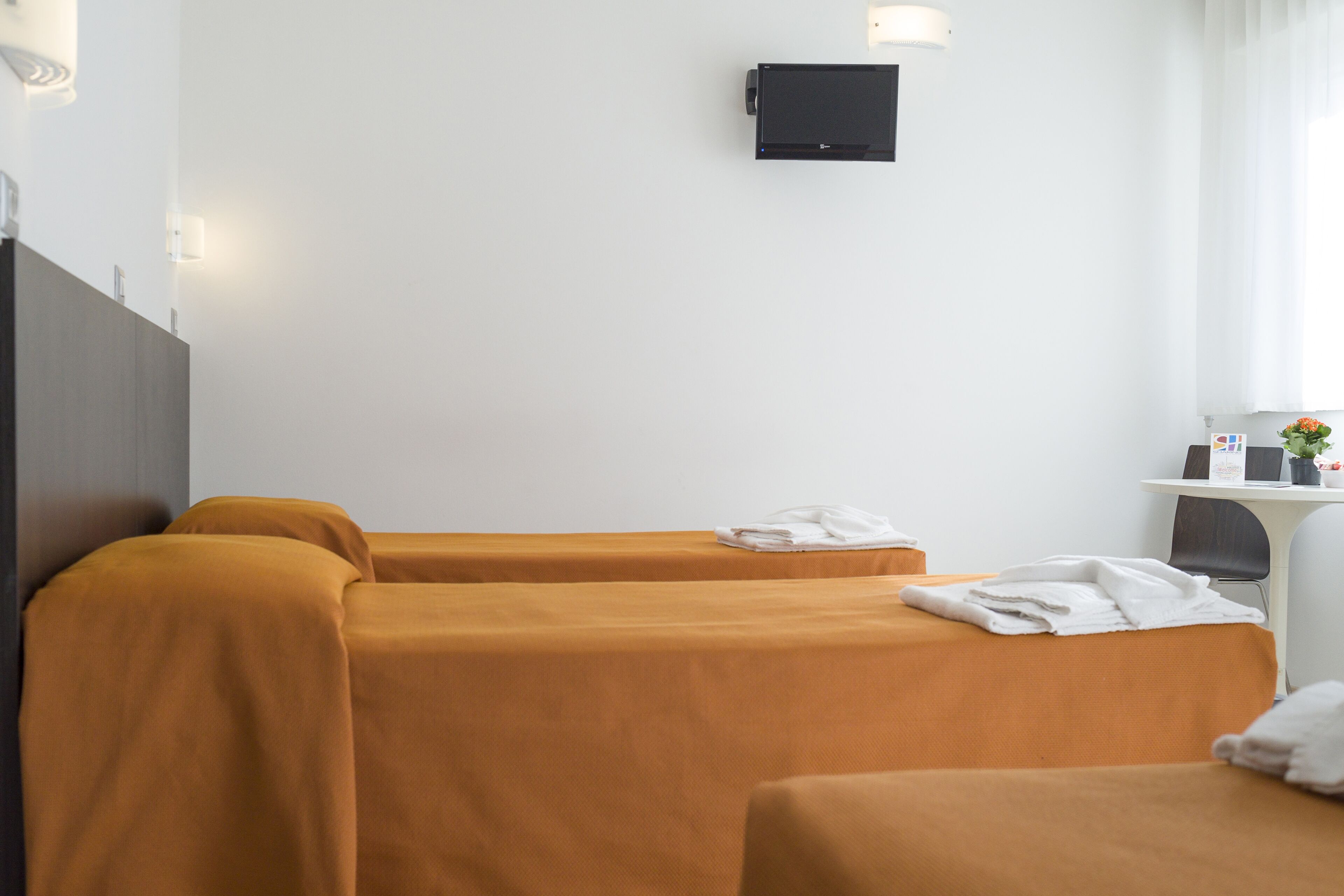 A hotel room with two beds covered in orange sheets, a small desk with a computer and a plant, and a wall-mounted TV.