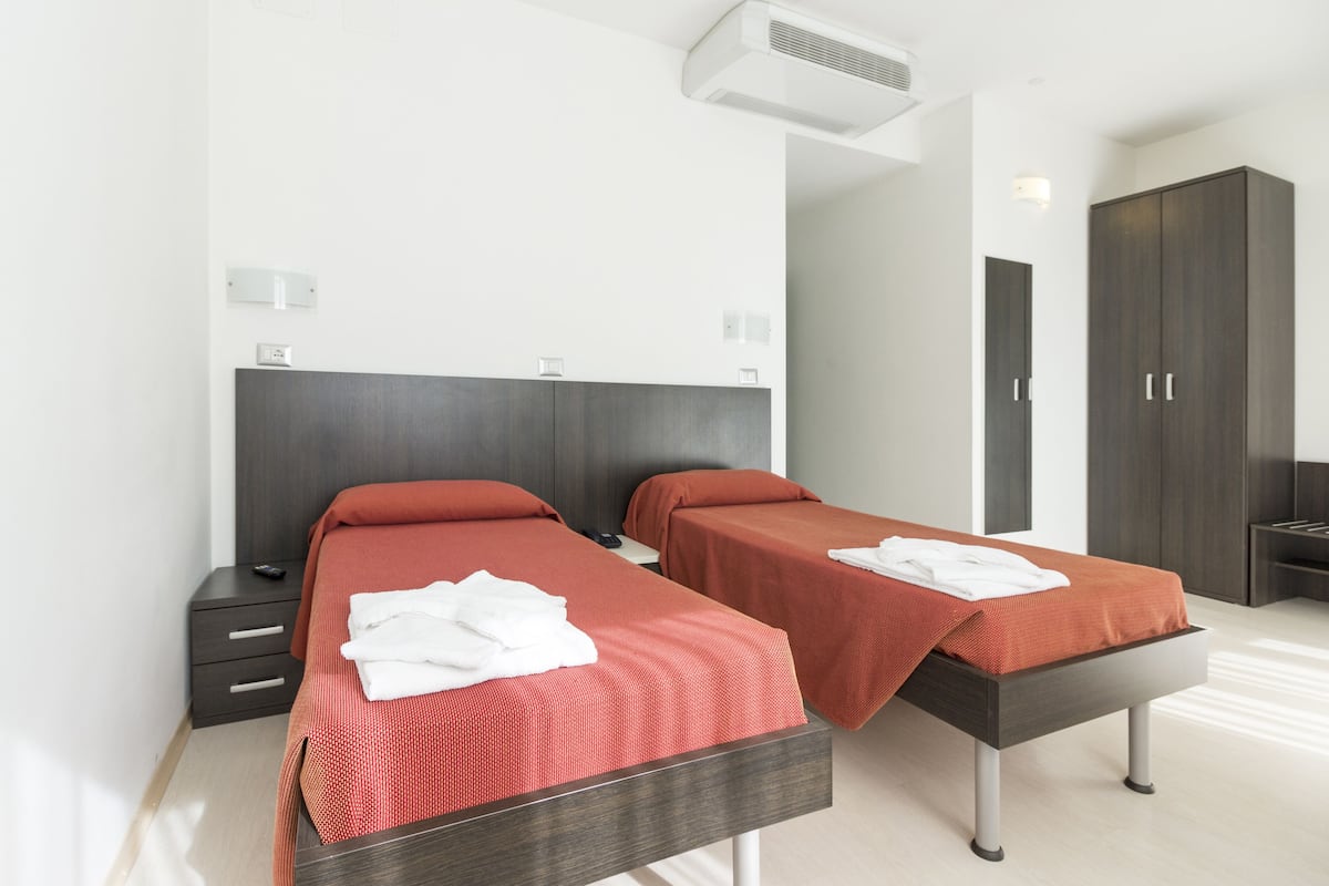 Hotellihuone, jossa on kaksi erillistä sänkyä, tumma puinen pääty, pieni yöpöytä, vaatekaappi ja kattoon asennettu ilmastointilaite.