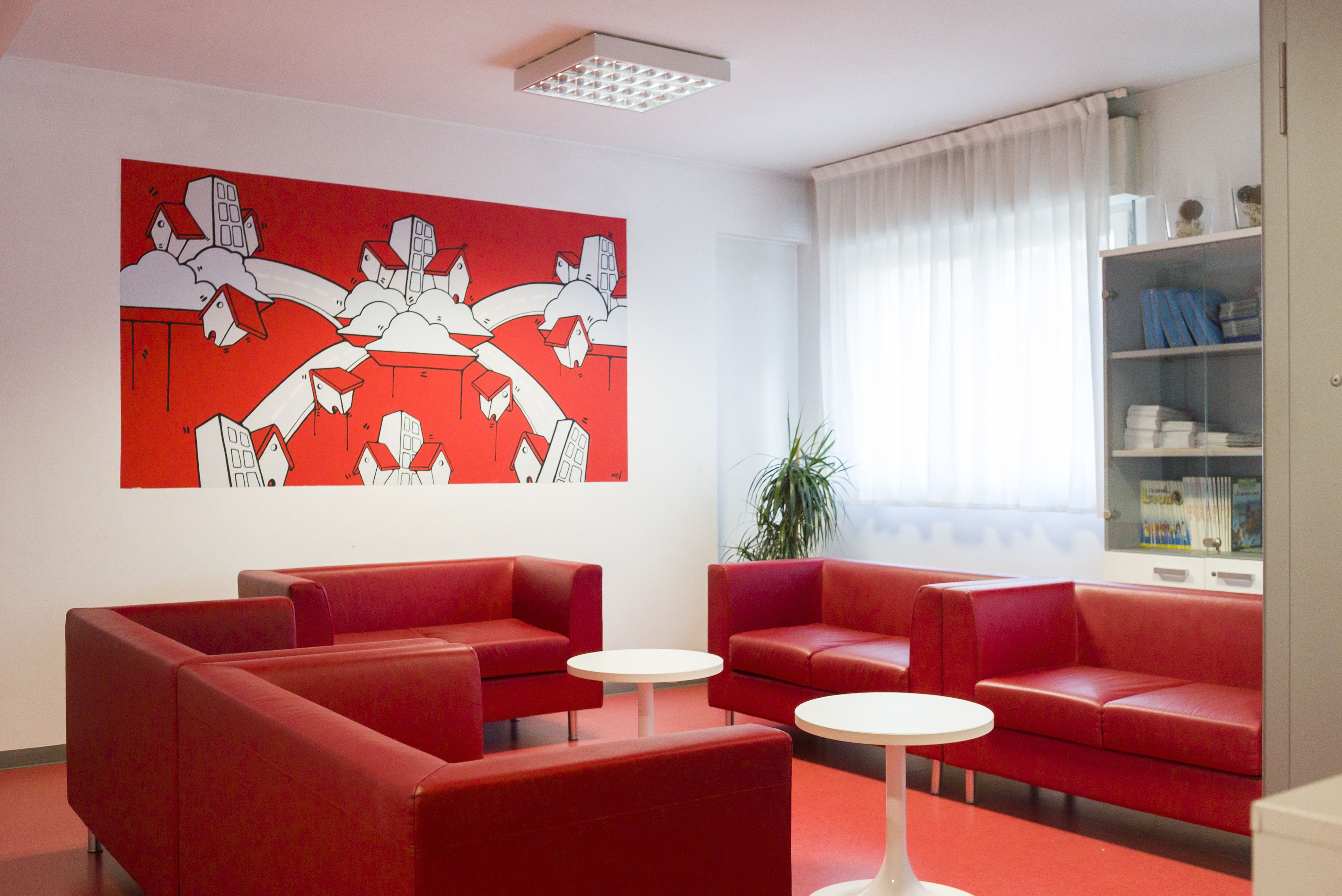 A red-themed waiting area with sofas and tables, a large abstract painting, and a bookshelf.