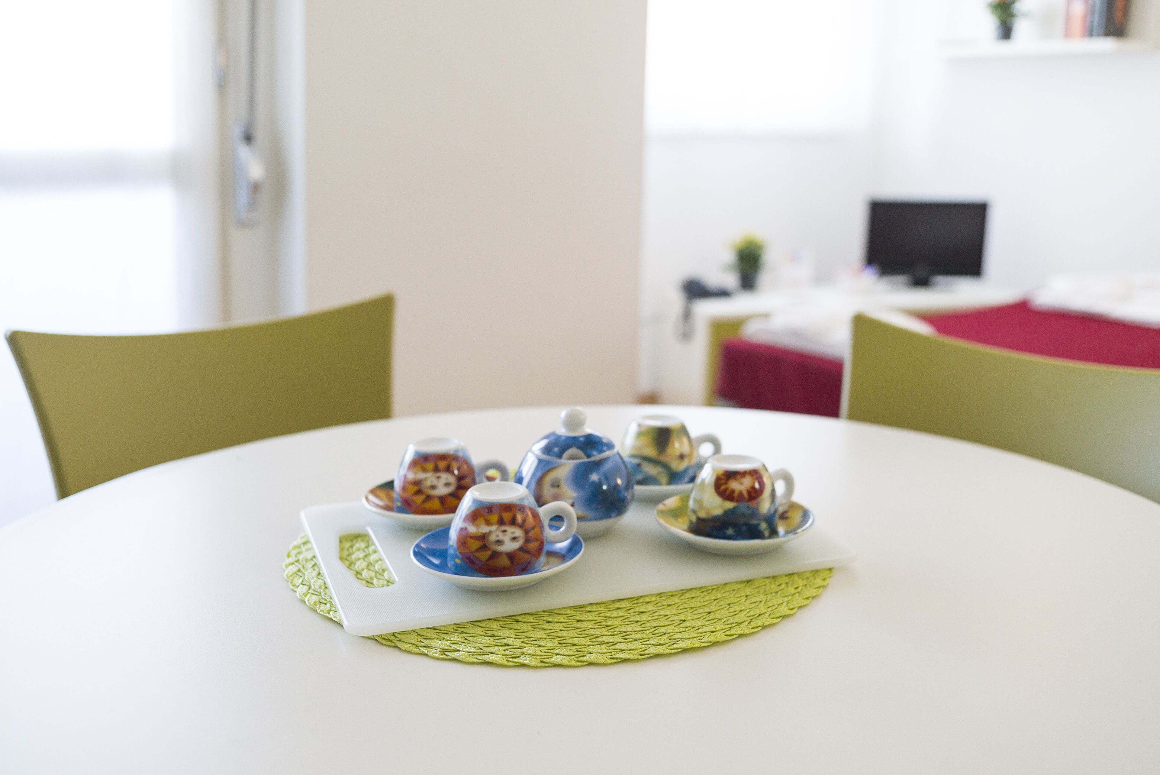 A round table with a woven placemat, surrounded by four ceramic cups with colorful designs, a white tablecloth, and a red table runner.