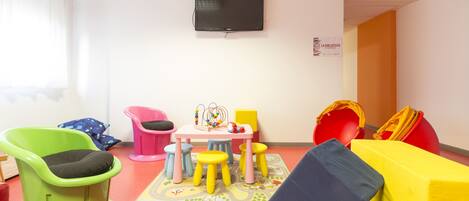 Zona de juegos infantiles con sillas de colores, una mesa pequeña y un televisor montado en la pared.