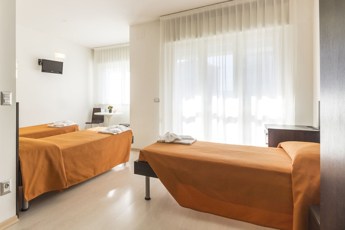 Hotellihuone, jossa on kaksi sänkyä, työpöytä, tuoli ja ikkuna, jossa on läpikuultavat verhot.