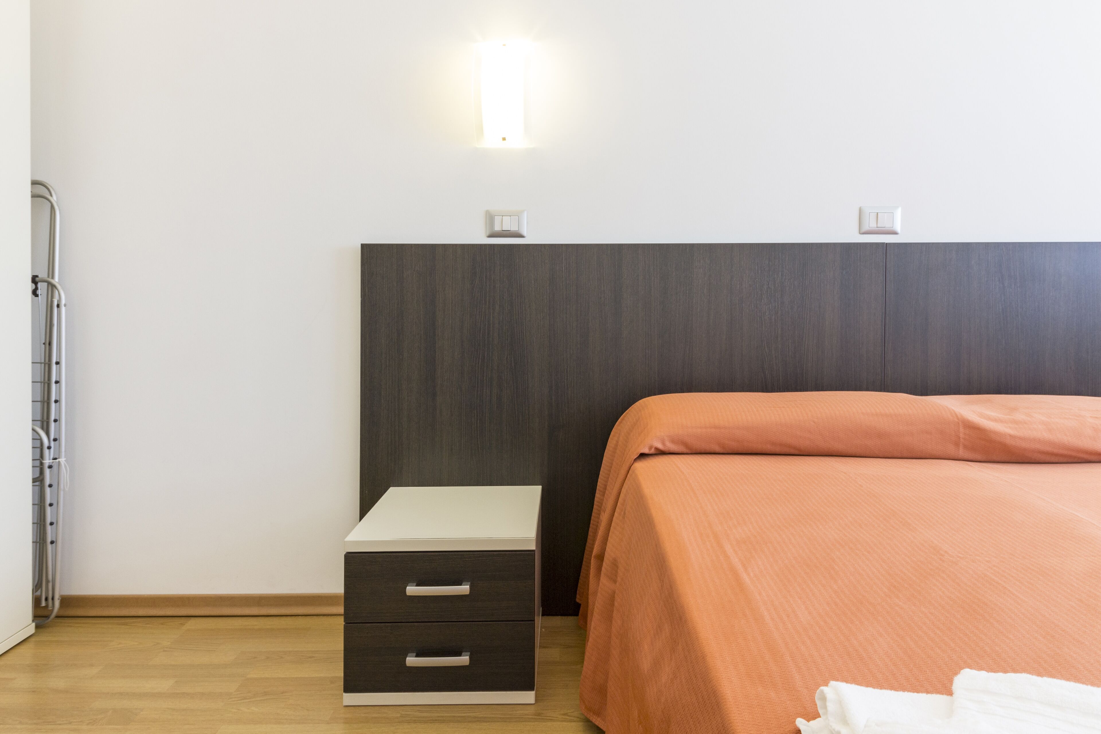 A bedroom with a bed, a nightstand with drawers, a wall-mounted light, and a radiator.