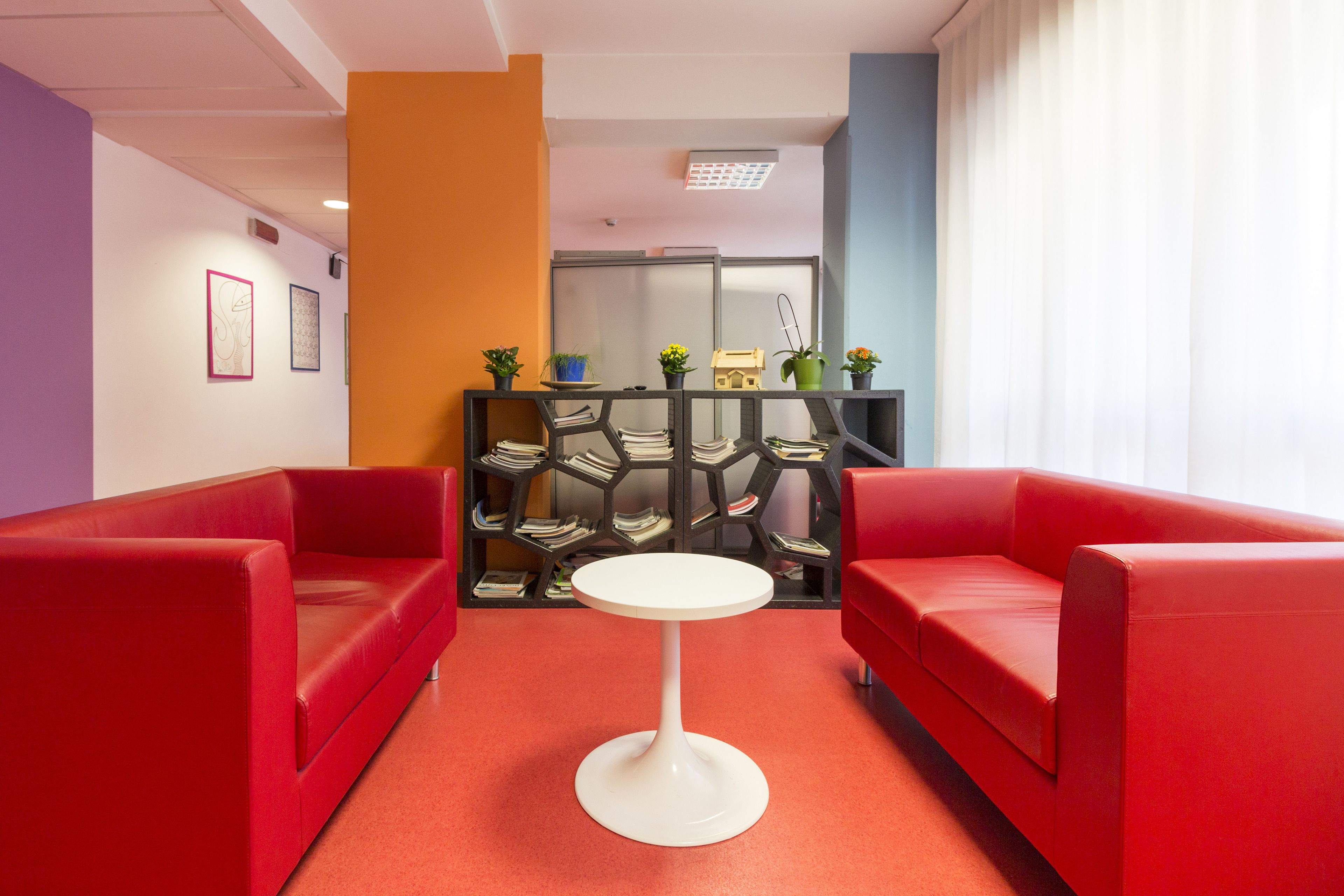 A modern lobby with red sofas, a white round table, and a bookshelf with magazines.