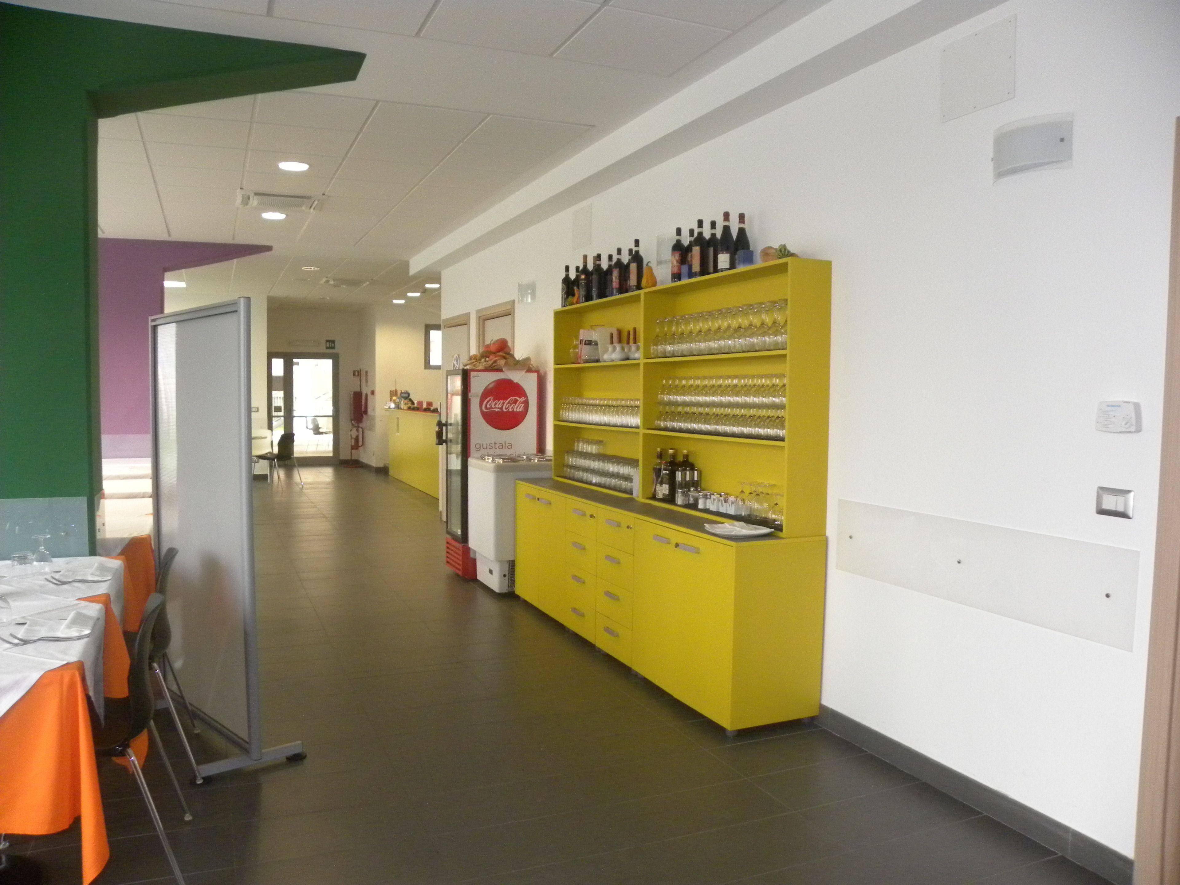 A hotel lobby with a Coca-Cola refrigerator, a yellow beverage station, and tables with orange tablecloths.