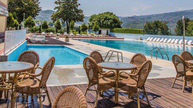 A pool area with lounge chairs and tables, surrounded by greenery and mountains in the background. - Hotel Columbano (Peso da Régua)