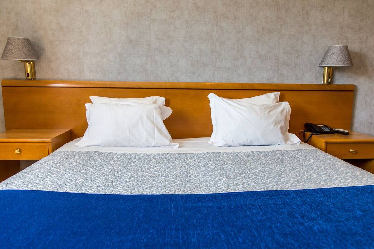 A hotel room with a wooden headboard, two white pillows, a blue and white patterned bedspread, and two bedside lamps.