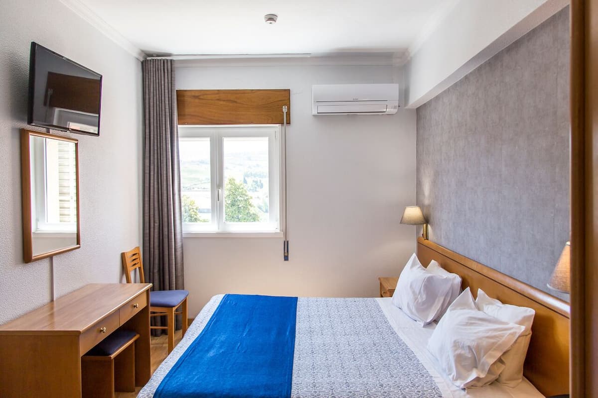 A hotel room with a bed, a desk, a chair, a TV, an air conditioner, and a window with curtains.