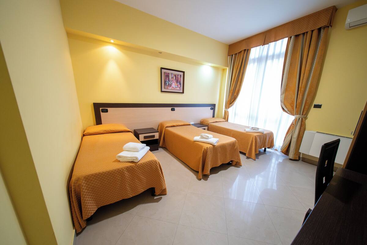 A hotel room with three beds, a desk, and a large window with curtains.