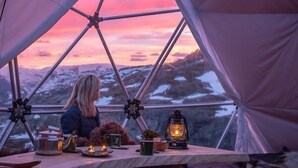 A geodesic dome tent with a view of a sunset. Inside, there is a table with a lantern, a kettle, and a fur rug. - Hordatun Hotel (Ullensvang)