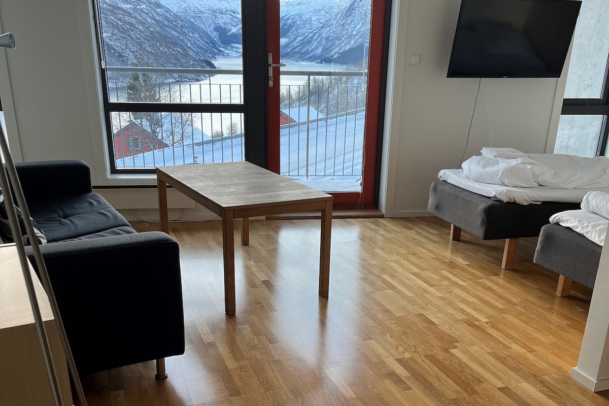 Uma sala de estar moderna com piso de madeira, um sofá, uma mesa e uma cama. Uma janela grande oferece vista para montanhas cobertas de neve.