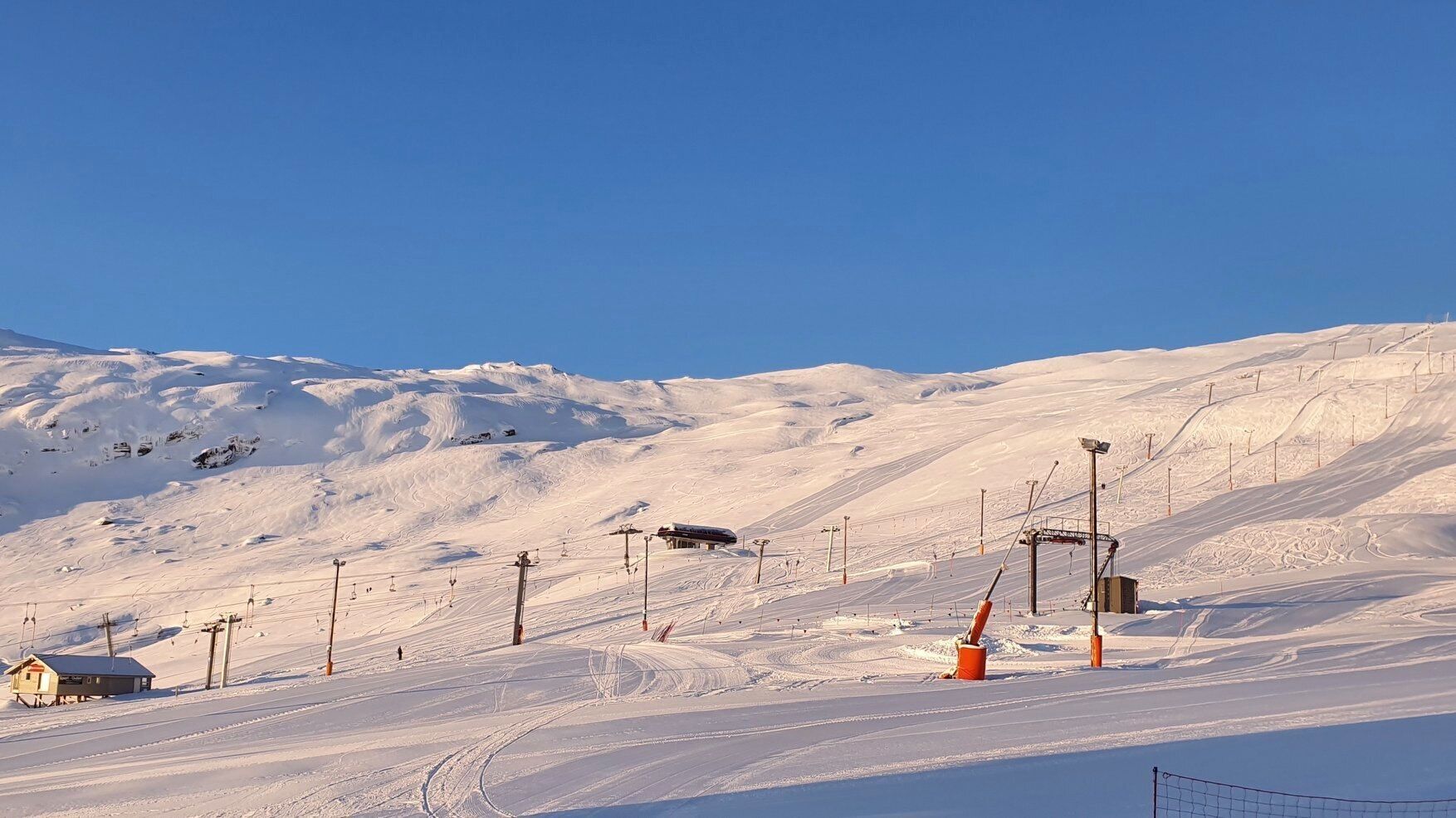 Pista de esquí cubierta de nieve con remontes, una cabaña pequeña y un cielo azul despejado.