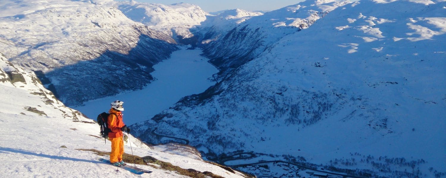 Una persona haciendo senderismo en una montaña nevada con un lago en el valle.