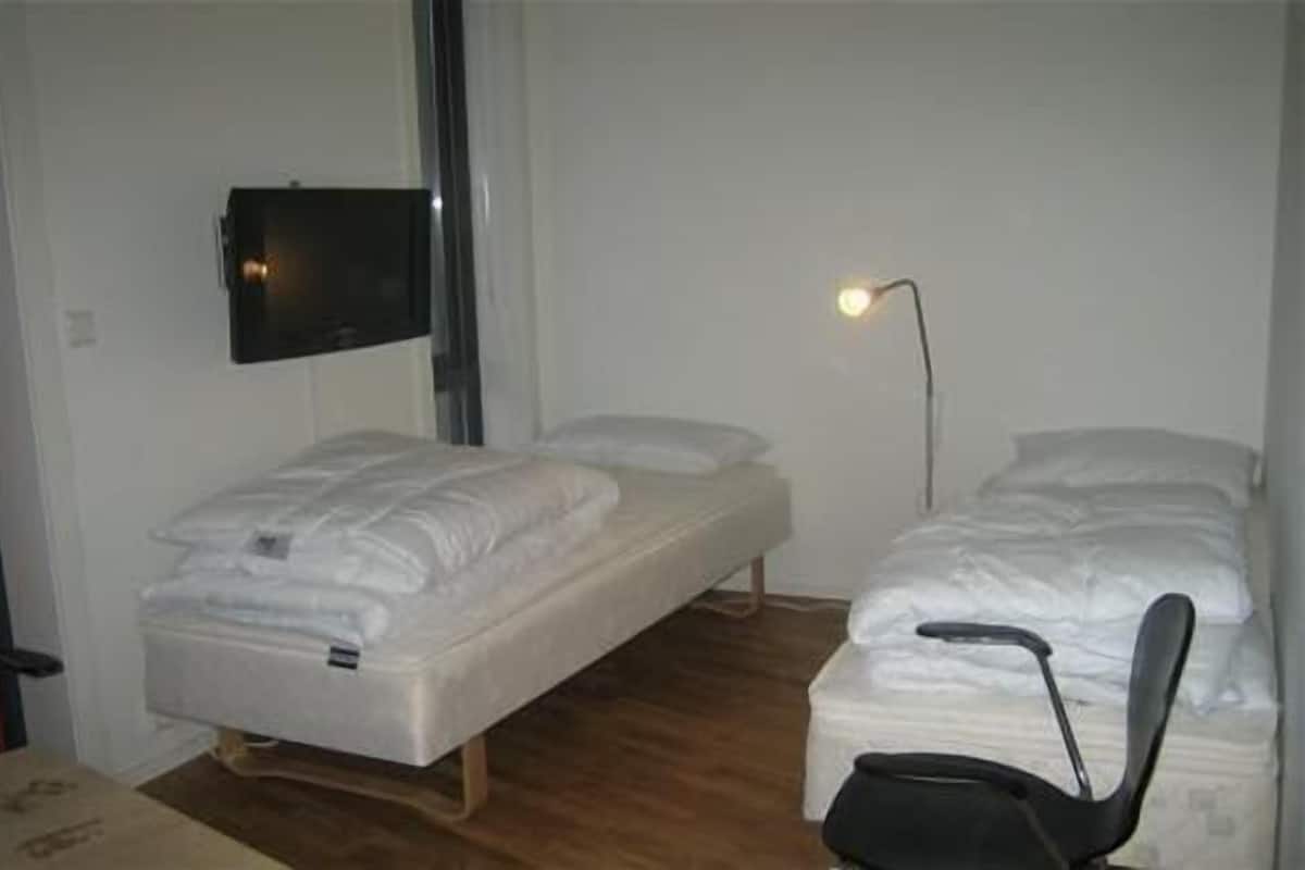 Quarto pequeno e pouco mobiliado com duas camas de solteiro, uma cadeira, televisão na parede e mesa de cabeceira.