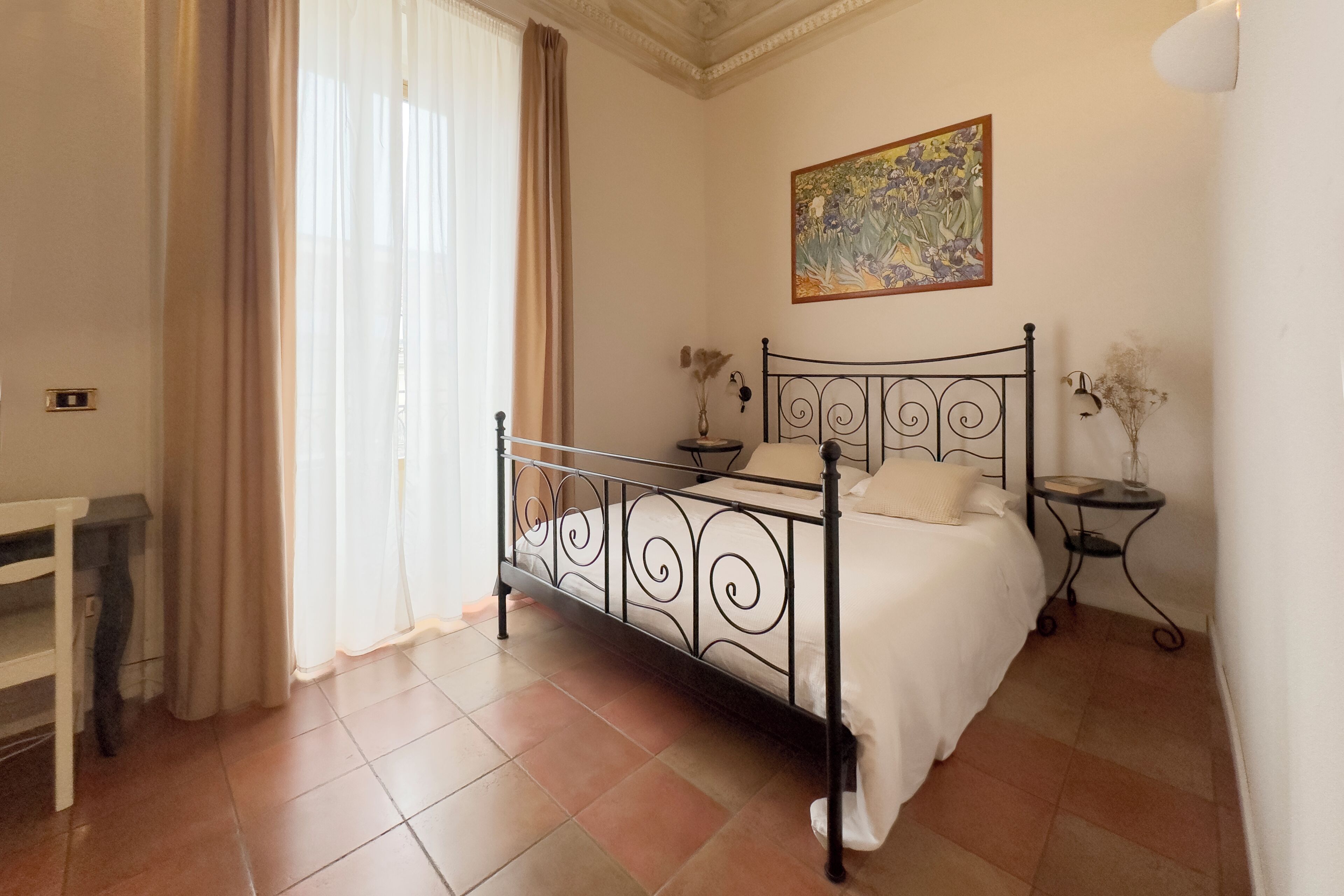 A bedroom with a bed, a nightstand, a small table, a chair, and a painting on the wall.