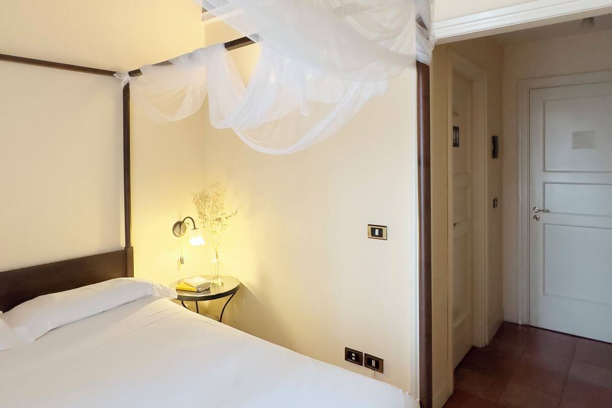 A four-poster bed with a canopy, a bedside table with a lamp, and a door in the background.