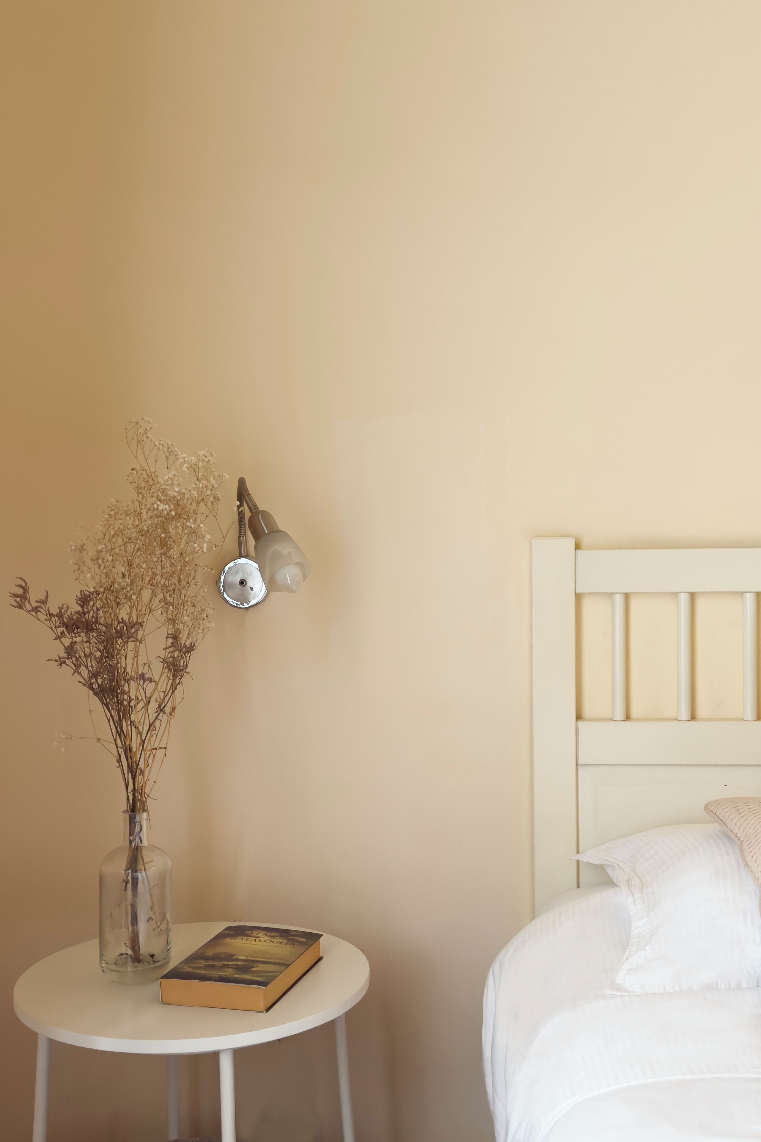A room with a bed, a small round table with a book and a vase of dried flowers, and a wall-mounted lamp.