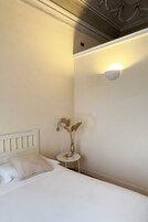 A neatly made bed with a white headboard, a small bedside table with a vase of dried flowers, and a wall-mounted light fixture.