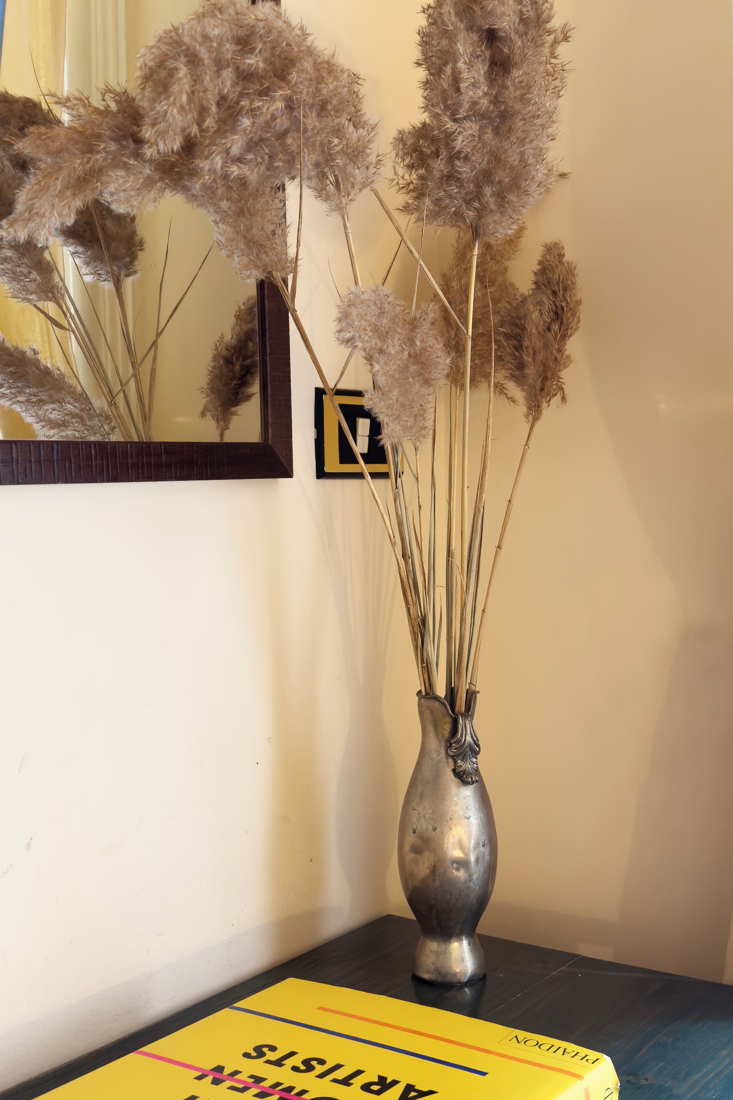 A vase with dried pampas grass on a table next to a book titled "ARTHUR'S MYSTERIES."
