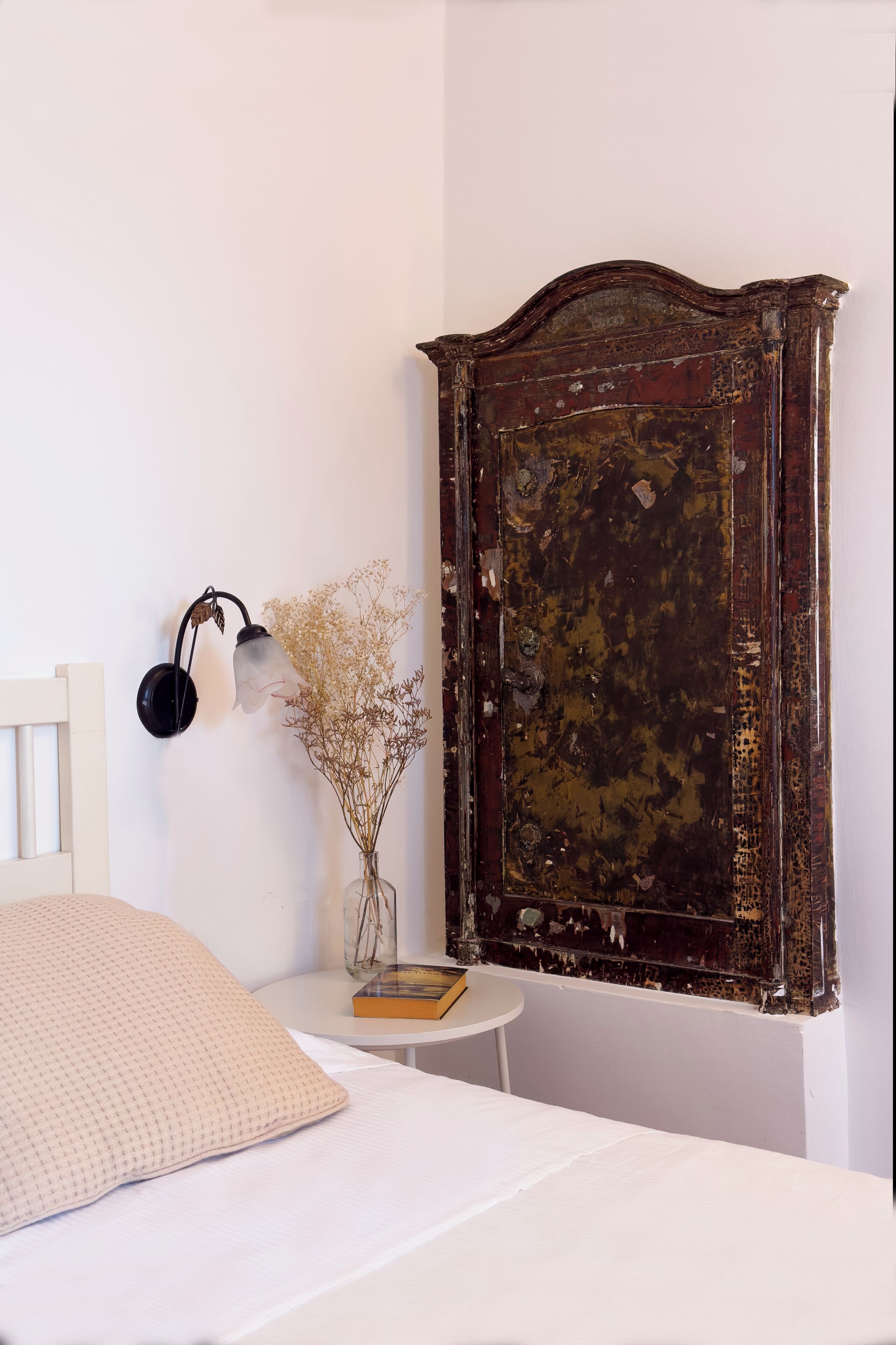 A bedroom with a white bed, a small white table with a book and dried flowers, a wall-mounted lamp, and an old wooden wardrobe.