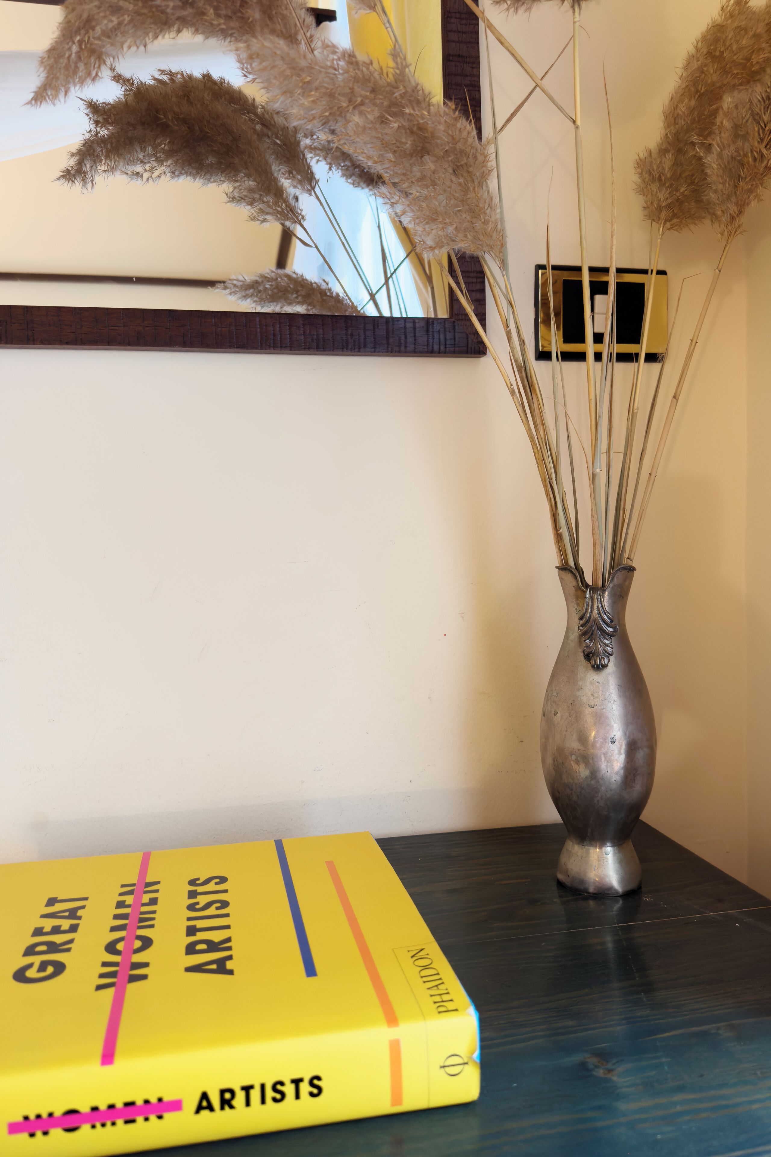 A book titled "Great Women Artists" on a table next to a vase with dried plants.